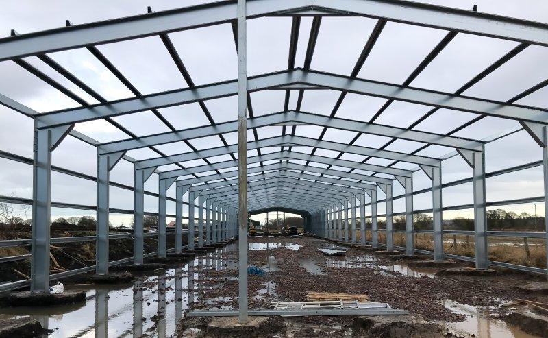 Interior view of a large steel building under construction, showing the skeletal grey metal framework, rafters, and support columns on a muddy site.