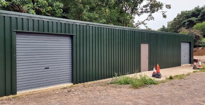 Side view of a long, dark green corrugated steel shed featuring a roller shutter door and a central personnel access door against a backdrop of trees.