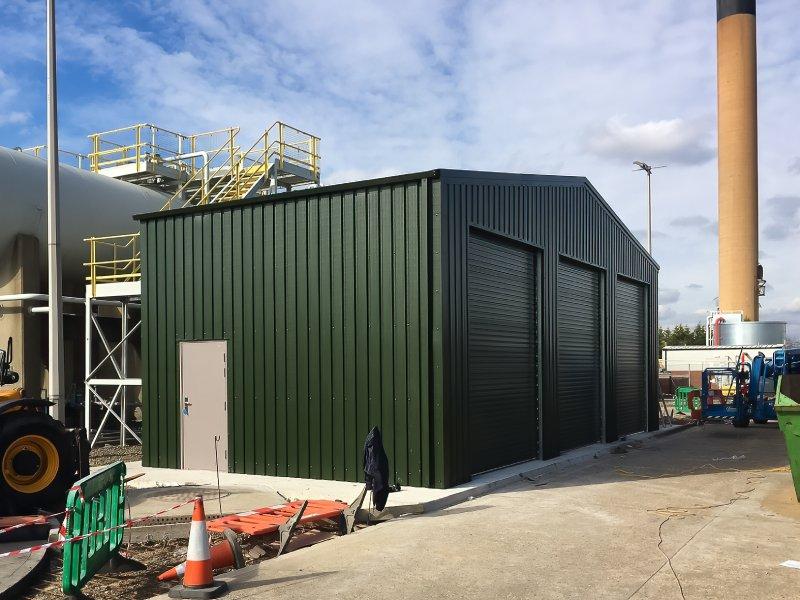 A dark green multi-bay steel workshop with three roller shutter doors and a side personnel door, located at an industrial facility.