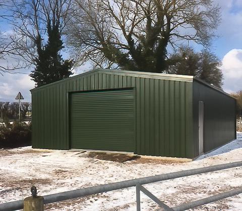 Exterior view of a large green steel portal frame building with a roller shutter door and side door in a winter landscape.